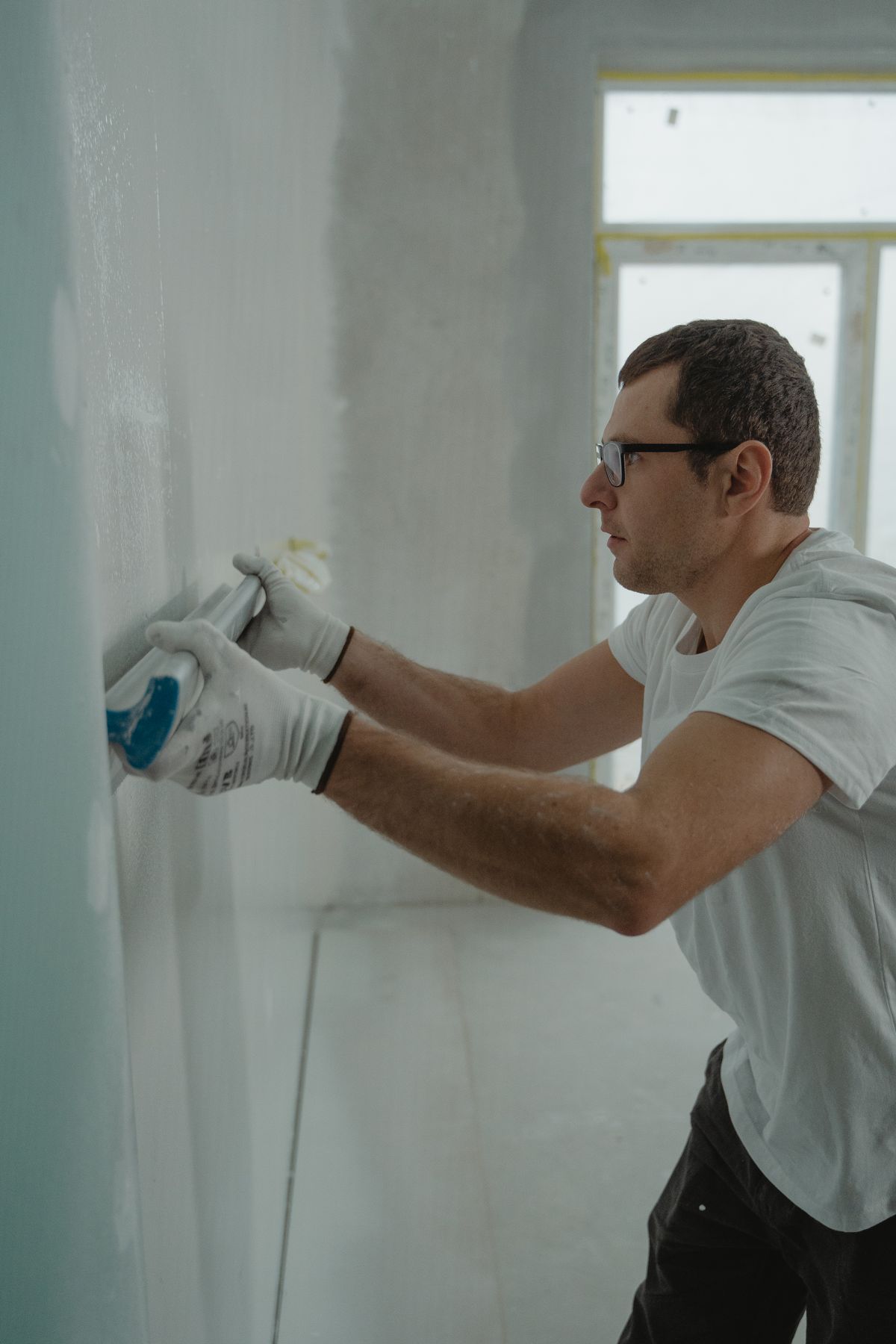 Finisher in work gloves smoothing joint compound along a drywall wall with a knife